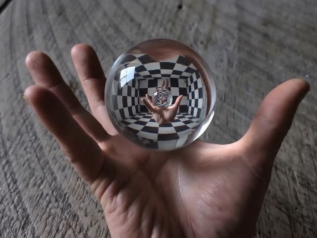 Una mano abierta sosteniendo una esfera de cristal transparente que refleja un patrón de ajedrez y un efecto óptico infinito de la misma mano.
